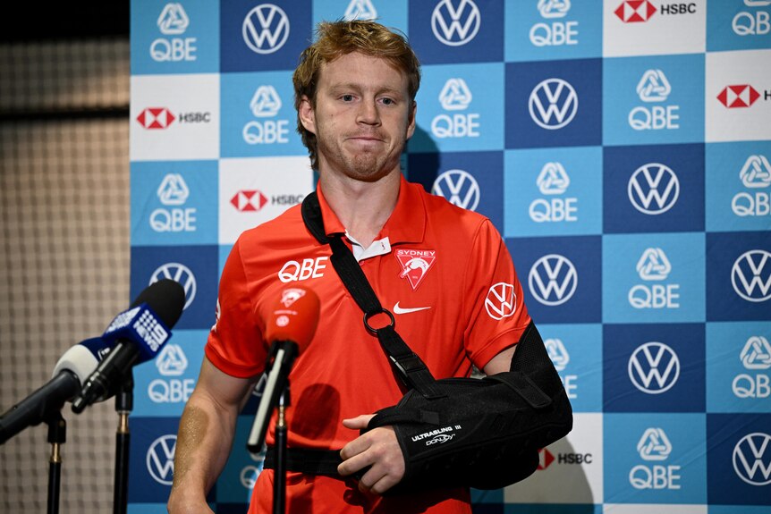 A dejected looking Sydney AFL player stands in front of microphones with his arm in a sling at a press conference.