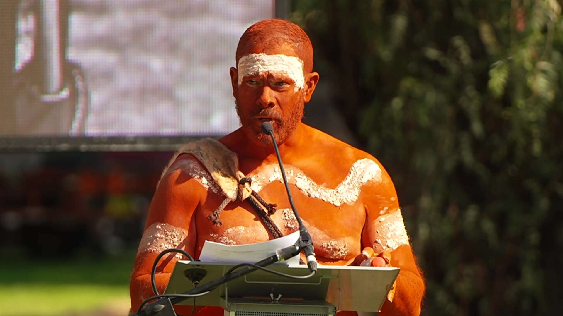 A man in Indigenous dance paint speaking at a lectern.