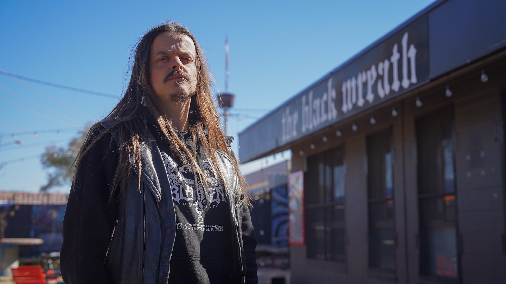 Pirate stands in front of The Black Wreath wearing a black leather vest over a black hoodie with his hair down past his shoulder