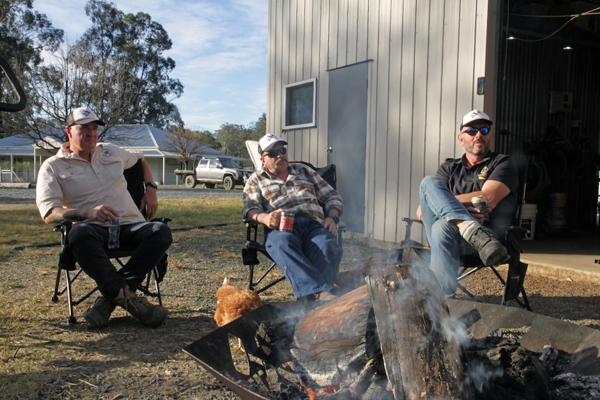 Three men sit around a smoldering fire during the day, all wear caps, two wear sun glasses, hold drinks, house in background.
