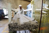 A factory worker wearing protective glasses and a cap moves steel rods at a manufacturing plant