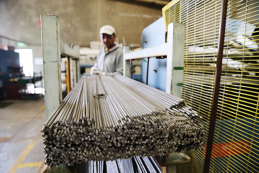 A factory worker wearing protective glasses and a cap moves steel rods at a manufacturing plant