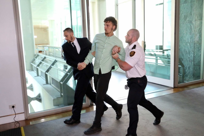 A protestor is escorted out of the public gallery by two security guards.