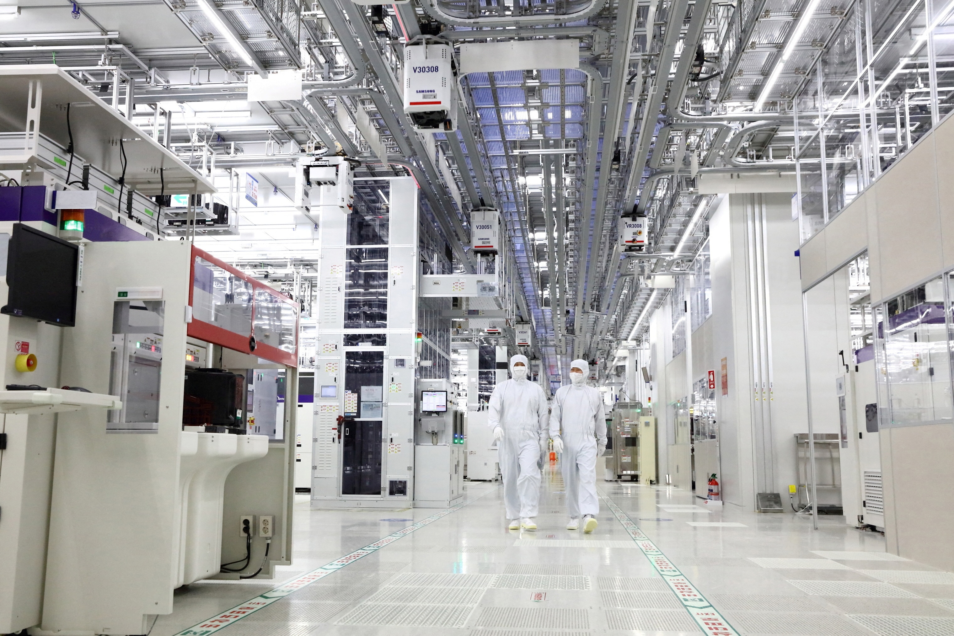 Two people in white coverall suits and masks walk through a factory full of beige electronic equipment.