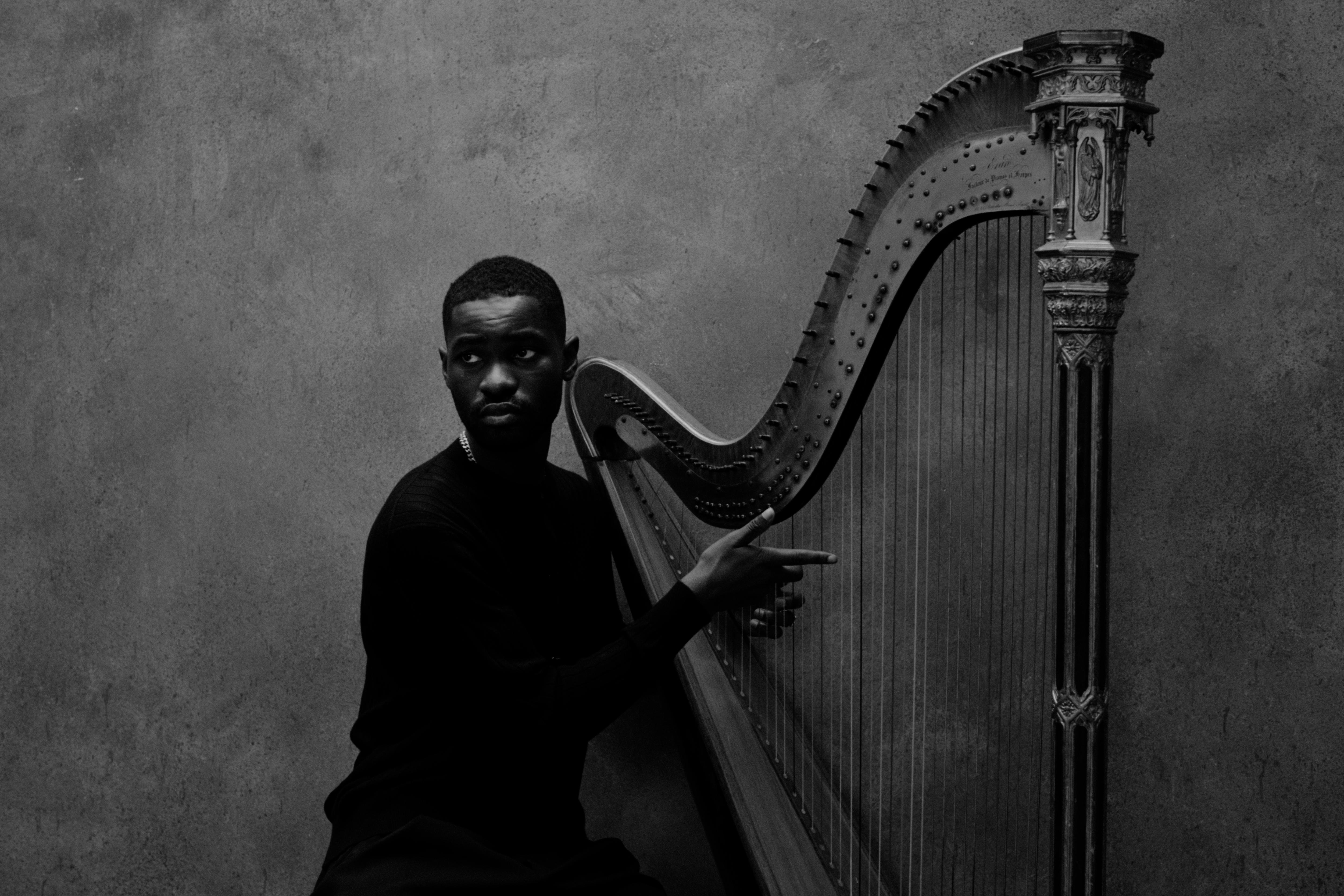 Darkened black and white photo of a man sitting at a harp with one hand on the strings as he looks to his right