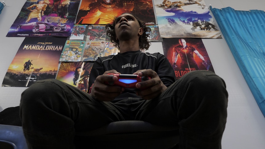 A low angle photo of a young Indigenous man holding a red controller and looking over the camera.