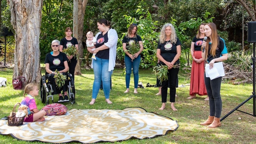 A group of women, a baby and young child stand around a circular mat on the grassed area, trees behind them, a mic and speaker.