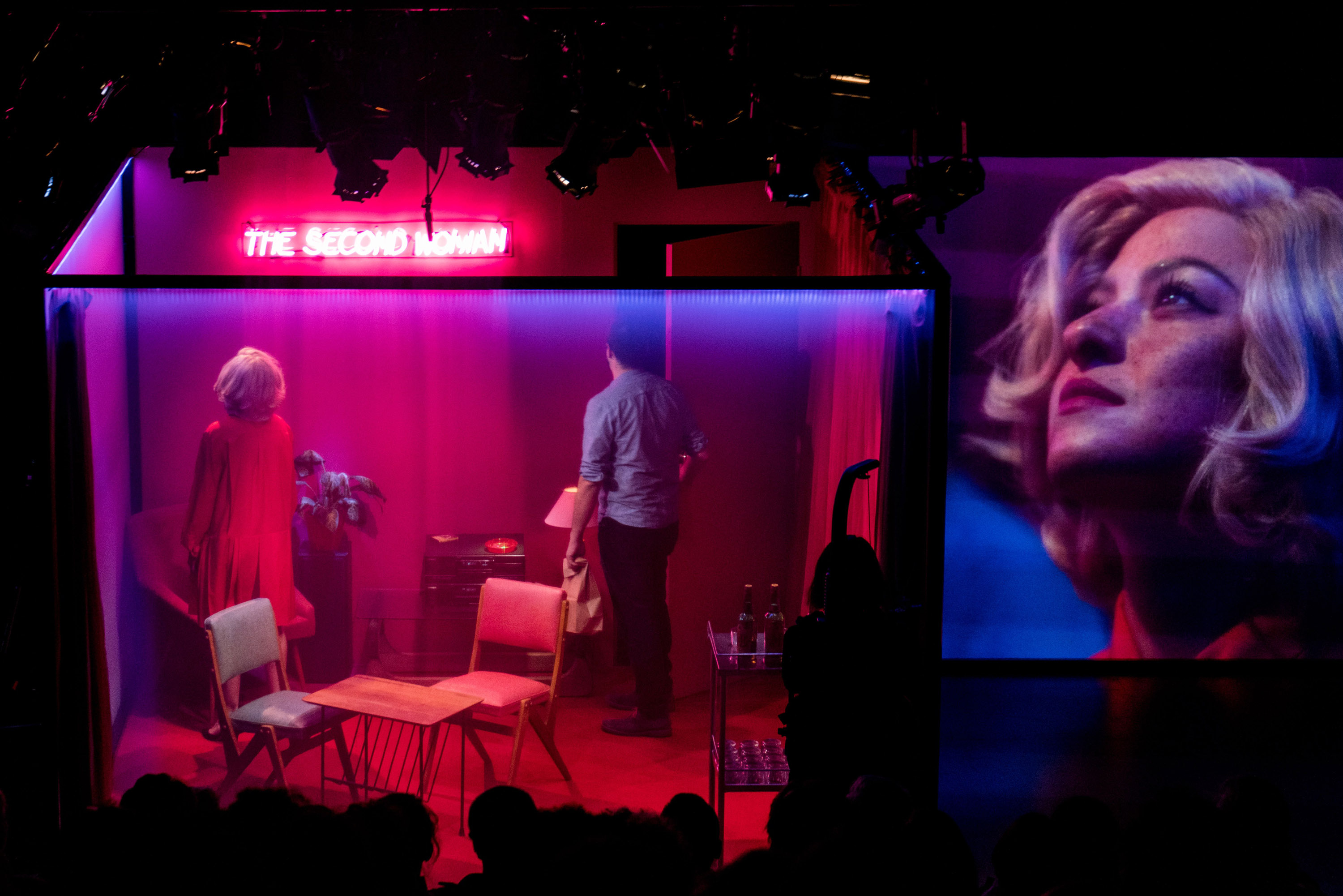 A woman stands in the corner of a set, facing away from a man opening a door. A close-up of her face is projected to a screen.