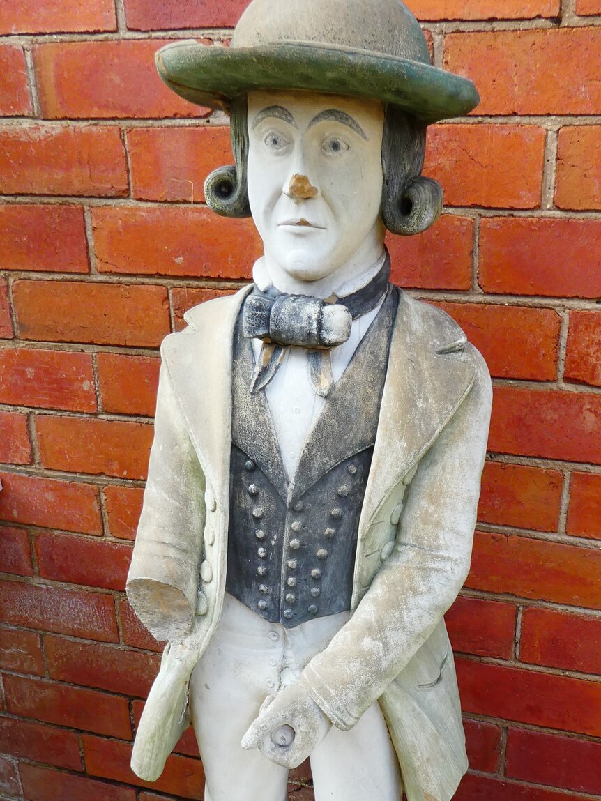 Statue of man wearing hat, long hair, well dressed in colonial clothing, holding penis.