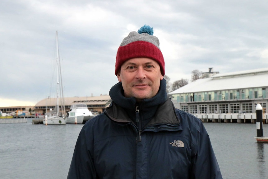 A middle aged man in warm clothes in front of a harbour filled with boats.