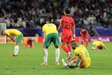 Soccer players wearing yellow and green sit in the grass while a player in red walks away after a game