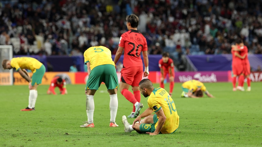 Soccer players wearing yellow and green sit in the grass while a player in red walks away after a game