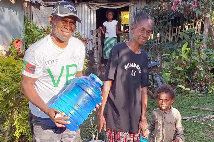 A smiling Vanuatuan man in a white T-shirt, holding a large water container, beside an older woman in blue t-shirt and child.