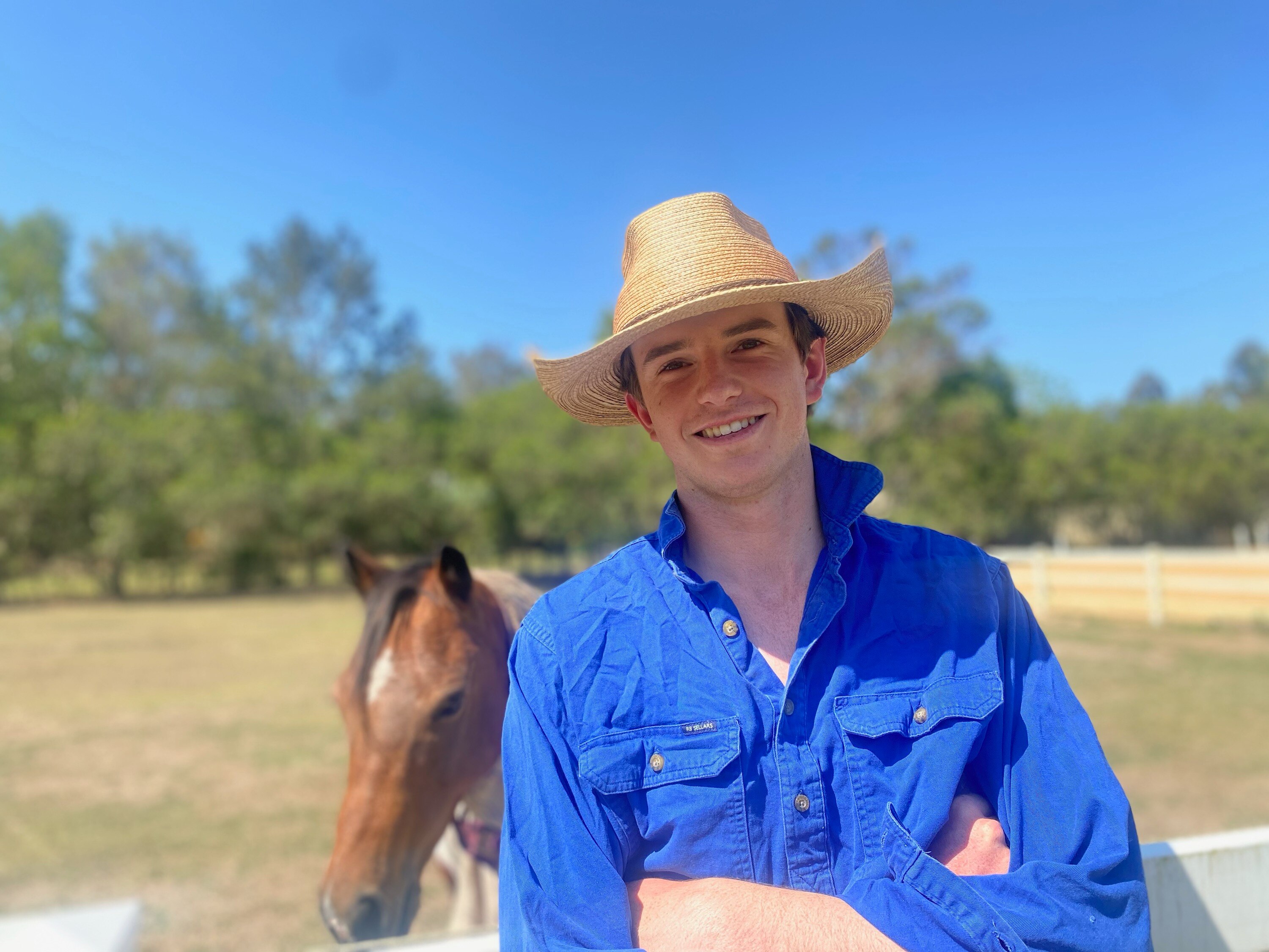 A young man with white skin smiles at the camera, arms crossed, in front of a paddock with a horse.