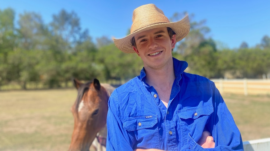 A young man with white skin smiles at the camera, arms crossed, in front of a paddock with a horse.