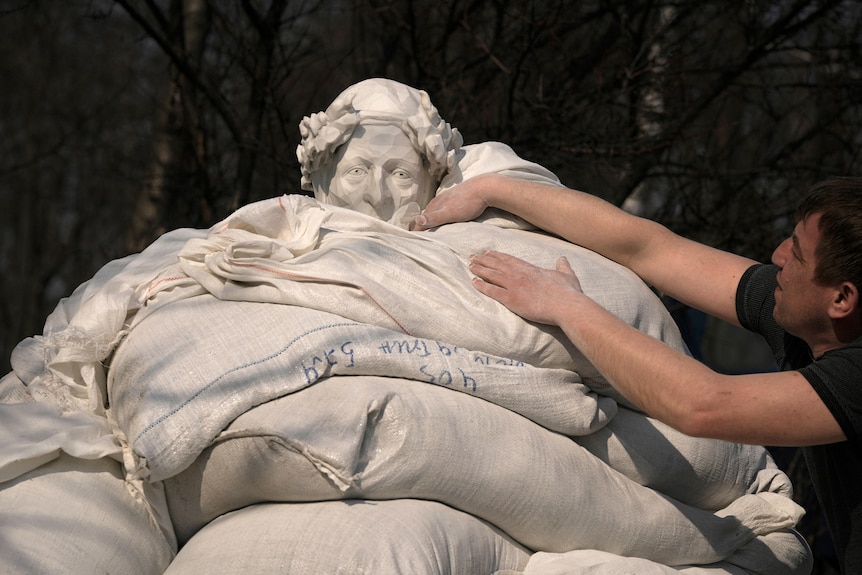 The top of a white statue is covered in sandbags - only the head is visible. A man reaches over to pat the bags around its neck.