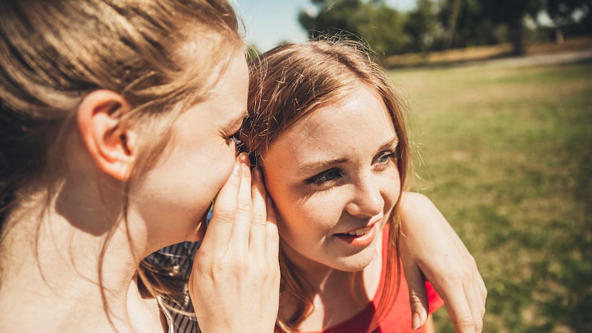 A young woman whispering in the ear of a female friend.