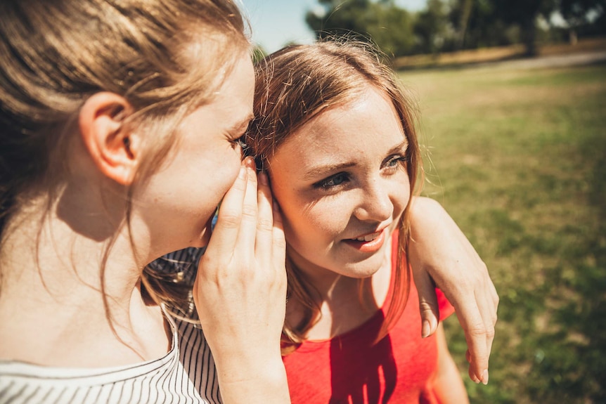 A young woman whispering in the ear of a female friend.