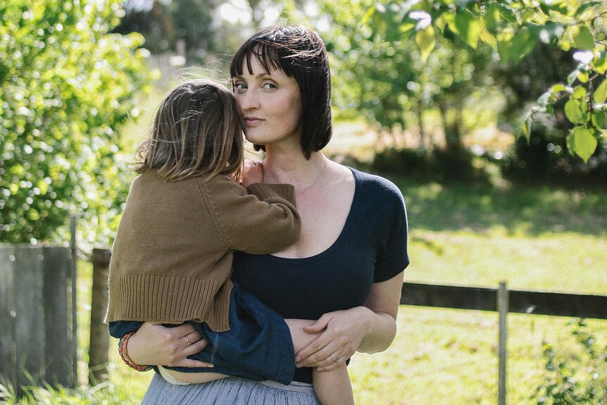 Jodi Wilson, with short black hair and neutral expression, stands holding small child, seen from the back, surrounded by trees..
