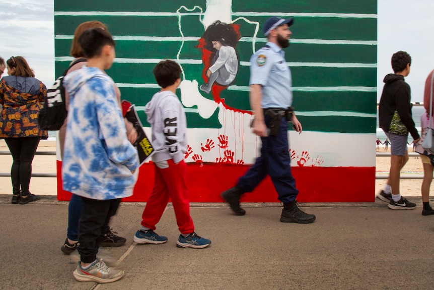 People of various ages, and a police officer, walk past a red, green and white sign, protesting against Iran