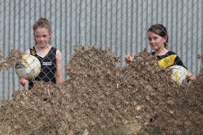 Two girls stand behind a fence filled with flood debris.