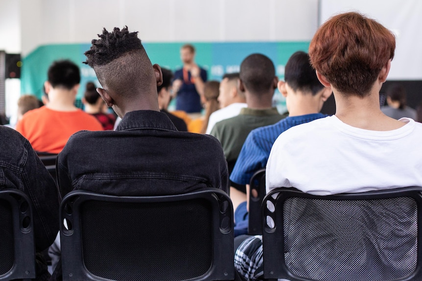 Students in a classroom with male teacher in background.