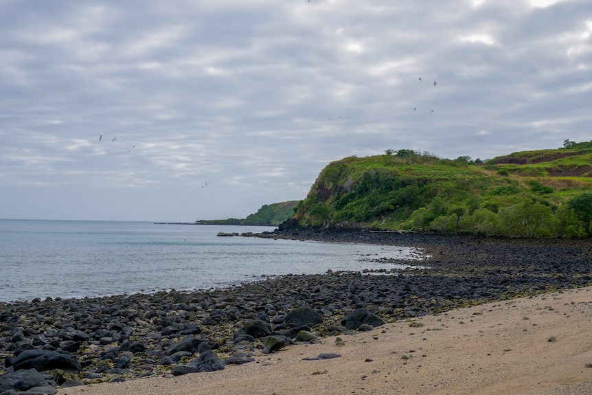 A sandy and rocky shoreline with green hills surround a bay.