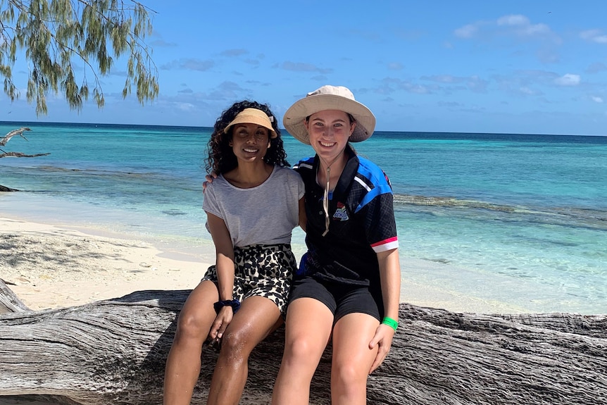 Two women sit on a log at a beach.