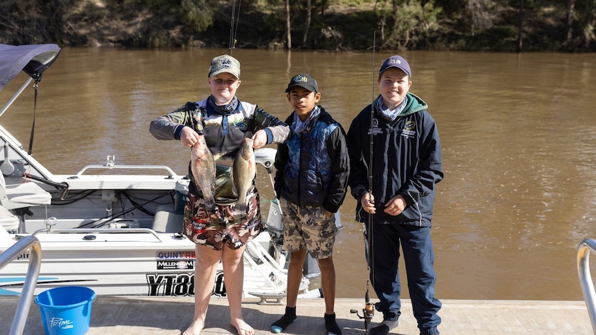 Three boys standing on a dock, with one holding two fish, and a brown river behind them.