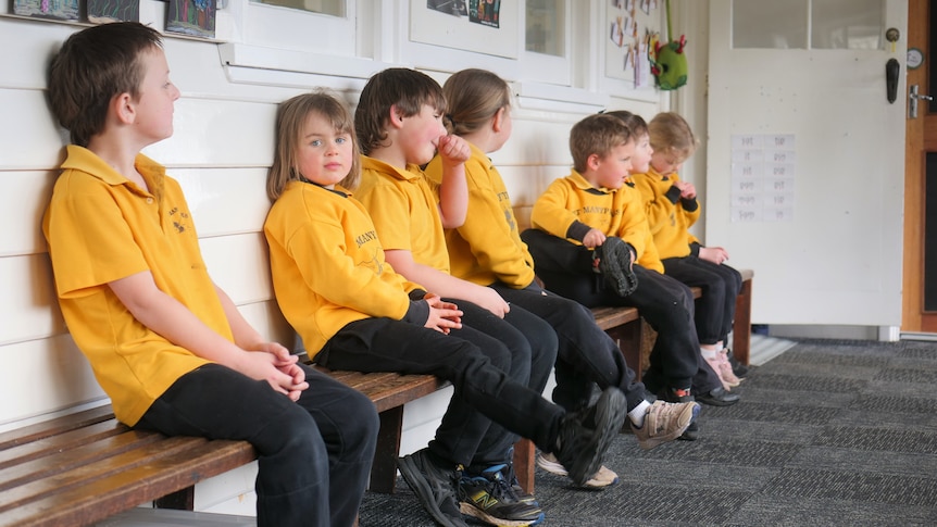 seven students in yellow t-shirt uniforms sit on bench outside classroom.