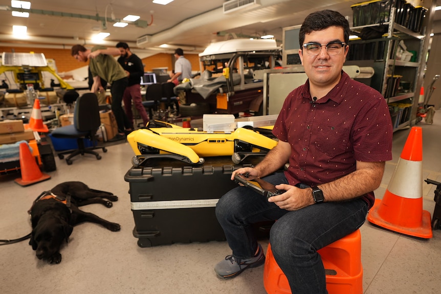 A man sits on a stool in front of a robotic dog with a guide dog lying next to it