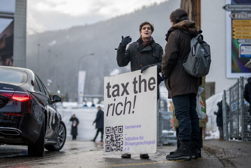 A woman stands on the side of a road holding a tax the rich sign.