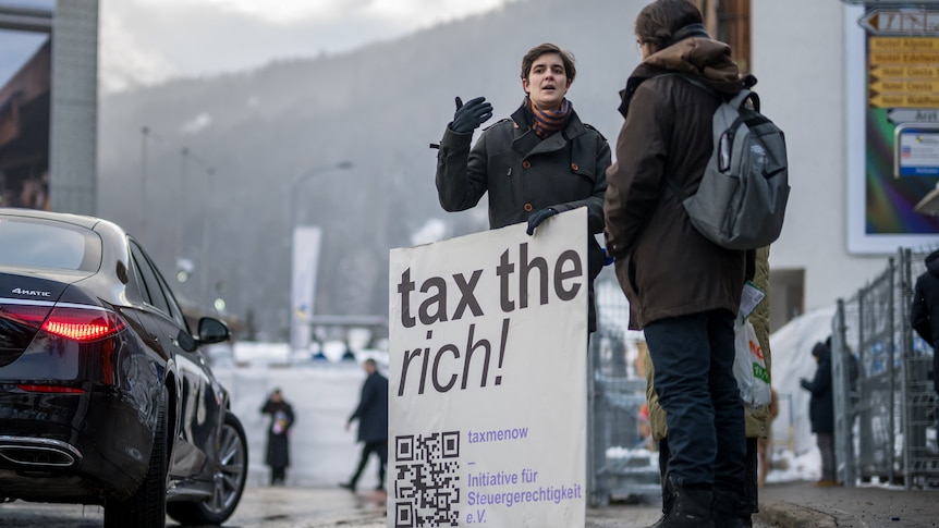 A woman stands on the side of a road holding a tax the rich sign.