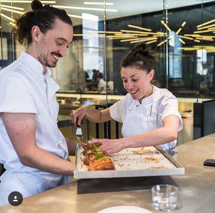 Man and woman in chef whites smiling big in bakery moving pastries on a board