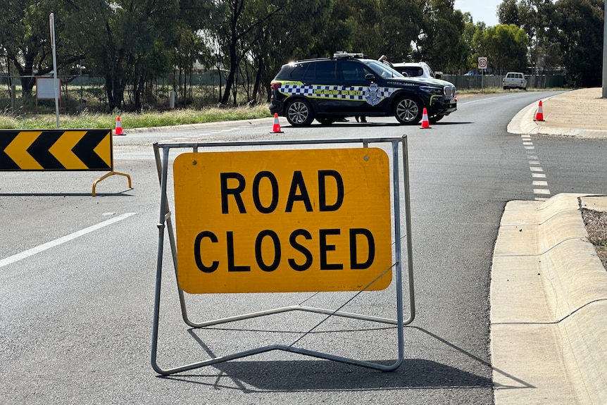 A yellow road closed sign stands on the roadway in front of a police car that is blocking the highway.