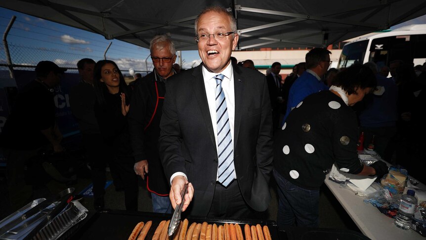 Wide shot of Scott Morrison standing in front of cooking sausages.