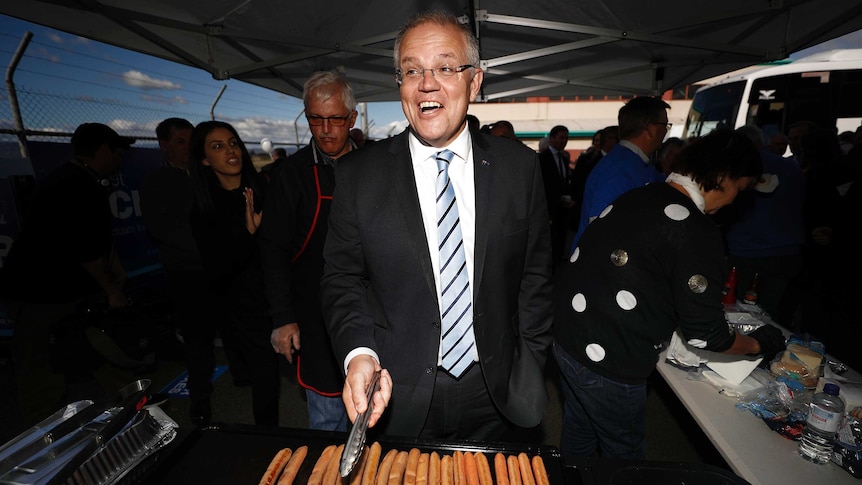 Wide shot of Scott Morrison standing in front of cooking sausages.