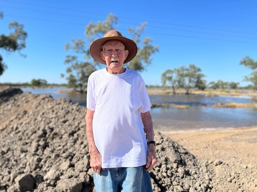 A man in a white shirt with a brown hat standing in front of a levee and water