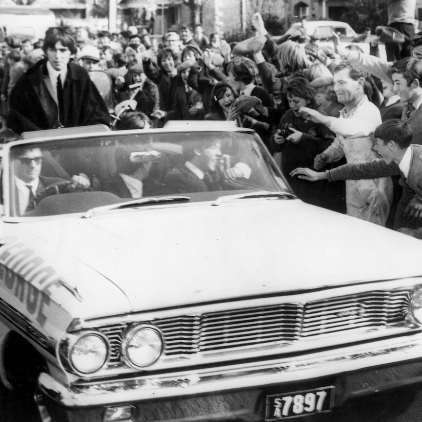 Black and white image of the Beatles driving in an open-top white car being mobbed by fans - adults and children. 