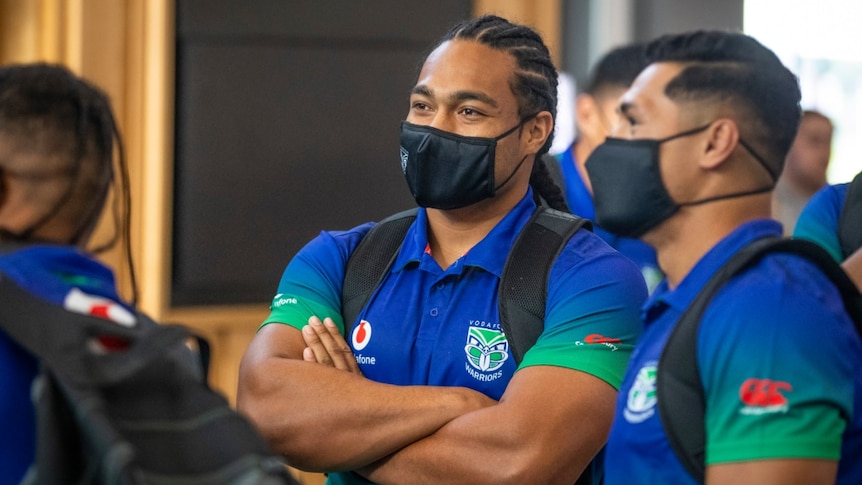 Two New Zealand Warriors NRL players stand next to each other with their arms folded wearing masks and backpacks