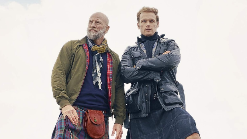 Two men, wearing tartan kilts, stand on a rocky outcropping on a green hill