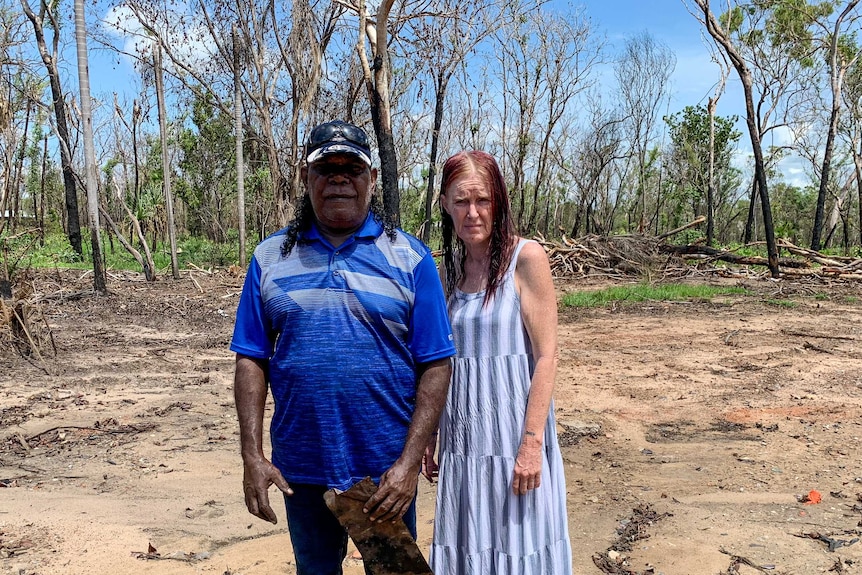 Jonathan Nadji in a blue polo shirt, standing with his partner in a striped dress, burnt tree trunk surround them.