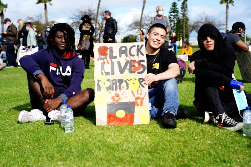 A group of rally attendees sit on lawn at Langley Park with a sign saying 'black lives matter'.