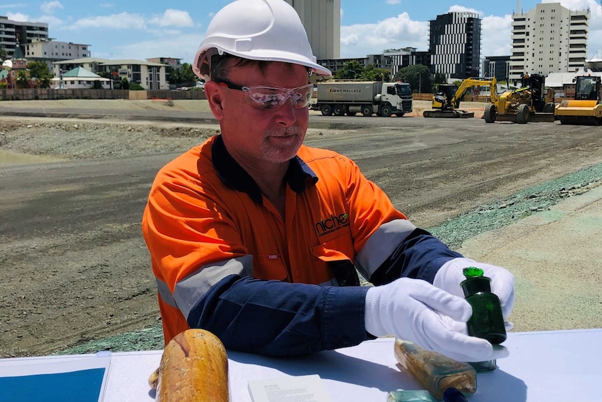 A man in a high viz work shirt and helmet holds a green perfume bottle while wearing white gloves