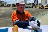 A man in a high viz work shirt and helmet holds a green perfume bottle while wearing white gloves