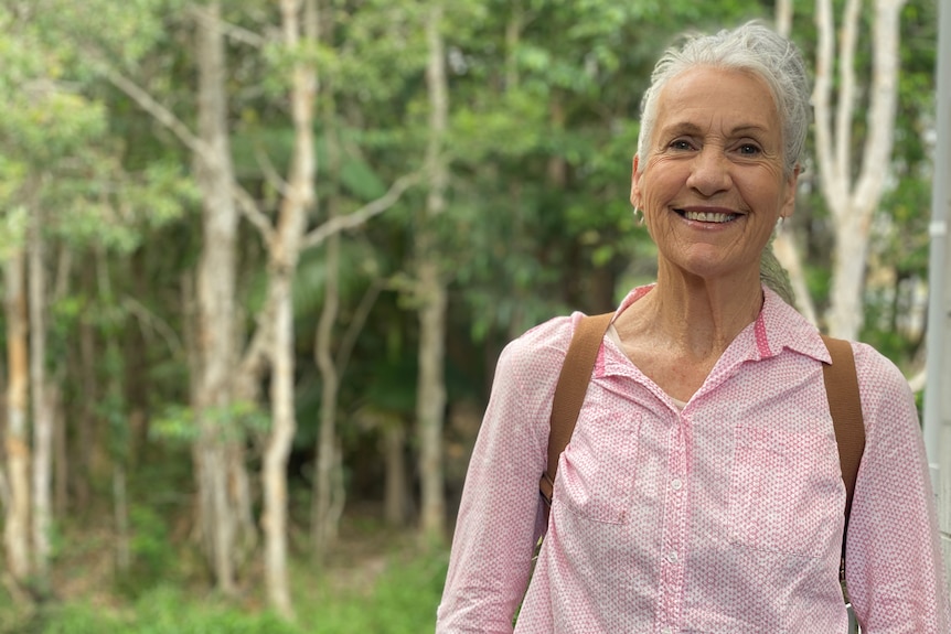 An adult woman in a pink shirt smiles at the camera while standing in front of trees.
