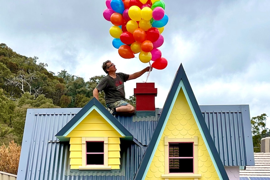 A man holds dozens of balloons over a huge, colourful wooden cubby house, as he sits on its roof.