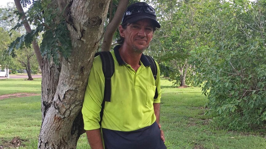 A homeless man in a hi-vis shirt and sunglasses stands next to a tree in a leafy-green Darwin suburb.