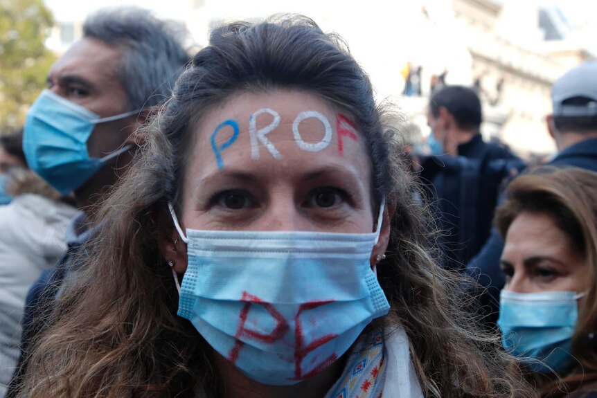 A woman with brown hair, wearing a mask, has the word 'teacher' written in French on her forehead.