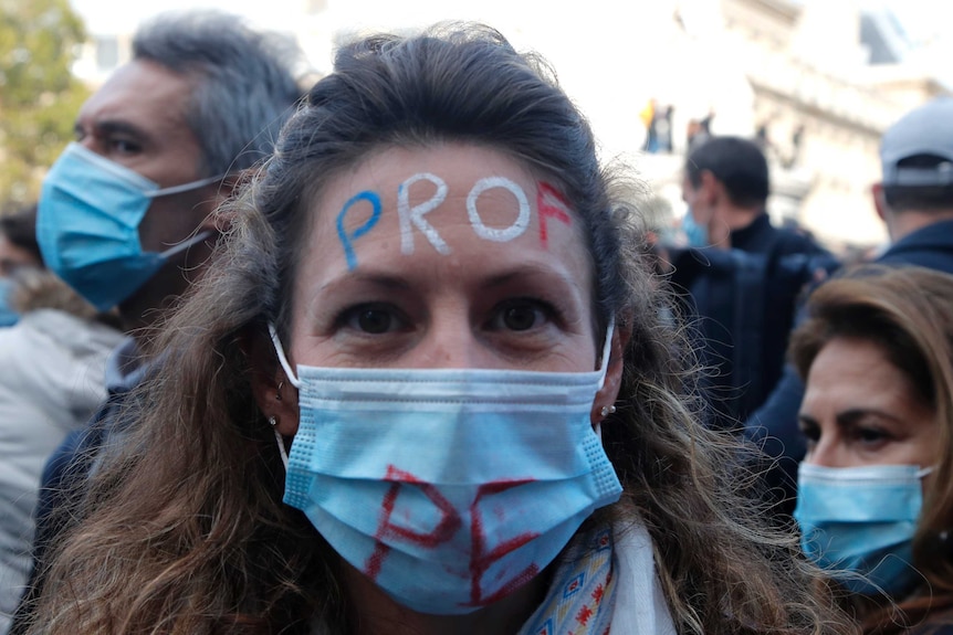 A woman with brown hair, wearing a mask, has the word 'teacher' written in French on her forehead.